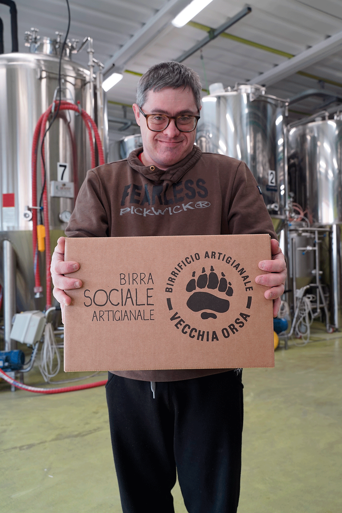 Un ragazzo sorridente con occhiali e felpa scura tiene in mano una scatola con il logo del birrificio artigianale "Vecchia Orsa Birra Sociale Artigianale"