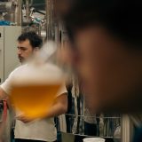 Un birraio in maglietta bianca osserva un bicchiere di birra in un birrificio, circondato da macchinari industriali. In primo piano, sfocato, un'altra persona e un bicchiere di birra